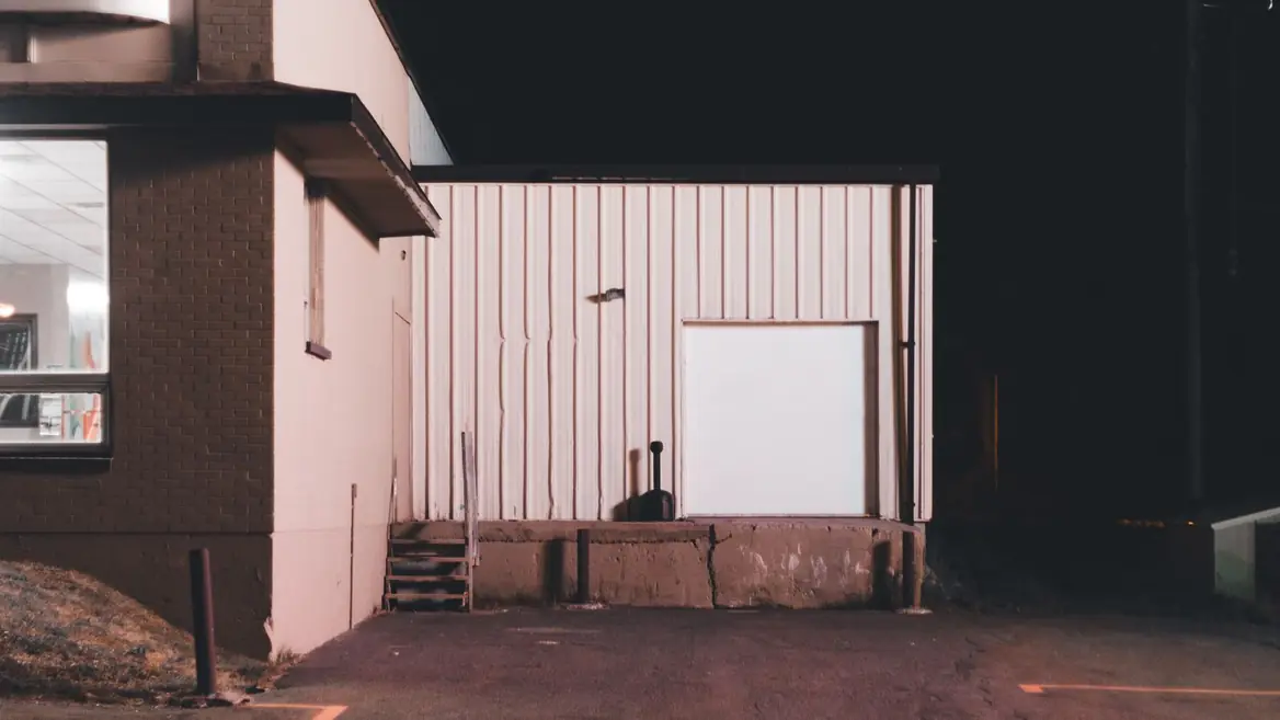 The rear of a small commercial building at night, one lit window and an empty lot.