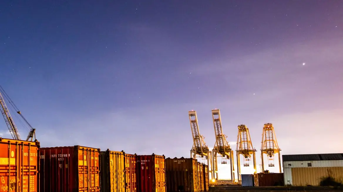 A container yard at dusk, stacked freight in the foreground and gantry cranes beyond it.