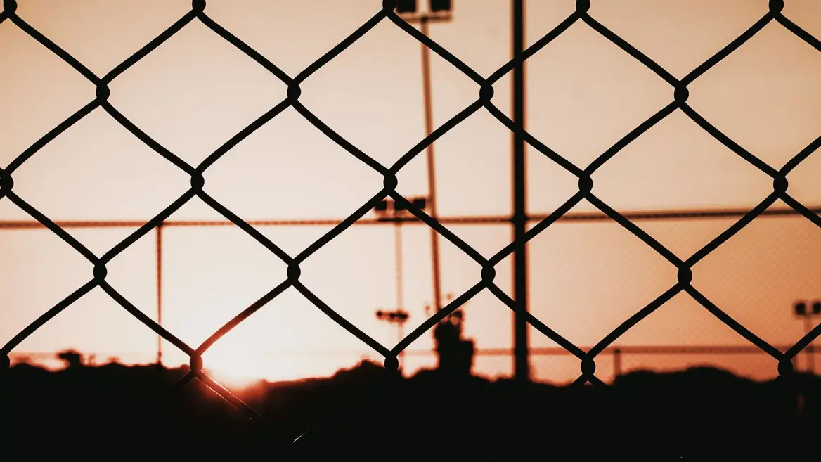 A chain-link perimeter fence in silhouette against a low sun, with floodlight masts beyond it.