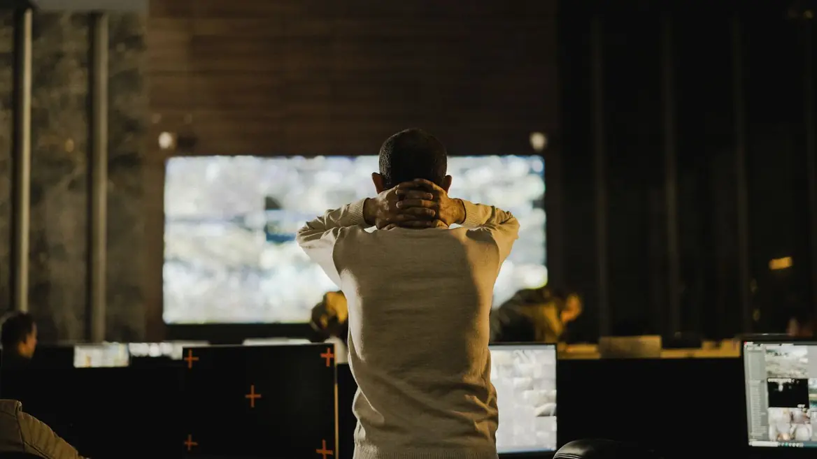 A person seen from behind in a darkened control room, facing a wall of camera feeds.